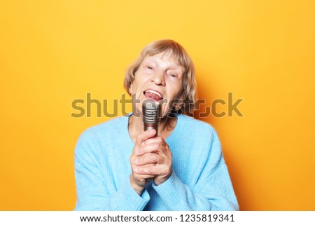 lifestyle  and people concept: Happy old senior woman singing with microphone, having fun, expressing musical talent over yellow background