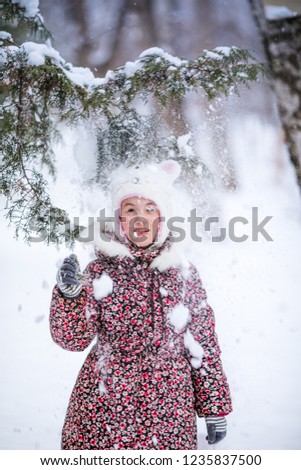 Smiling girl with white fur hat like a cat playing with snow. Winter snowy background and gteen trees.