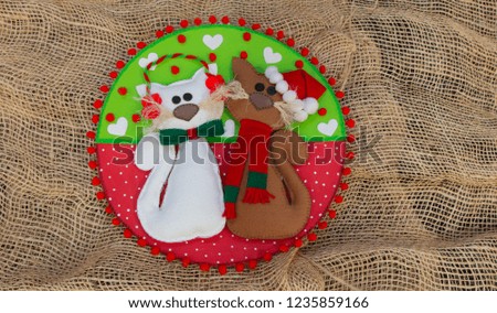 Pair of cats made in foam with Christmas decoration on a circle of red and green with white hearts, on a rustic background of jute fabric