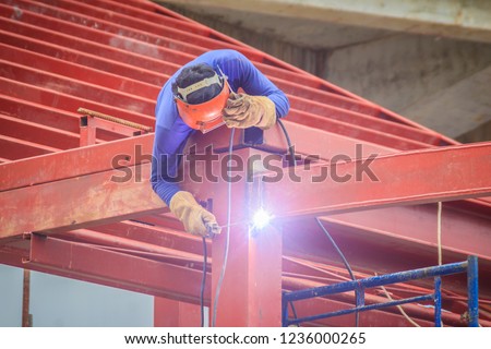 Risky welder while climbing and welding on top of the steel roof structure work at the building construction site. Skilled worker is welding on the high steel structure at the construction project. Royalty-Free Stock Photo #1236000265