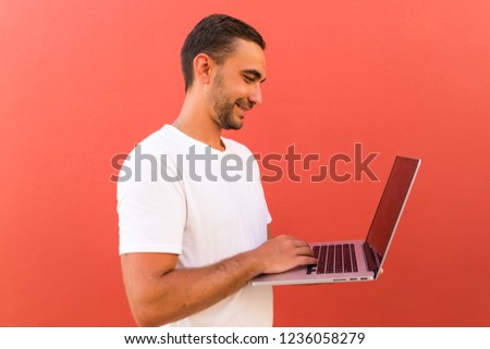 Picture of handsome man in casual t-shirt holding silver notebook and chatting or working isolated over red background