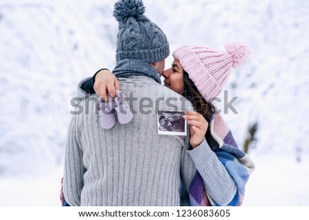 Beautiful pregnant woman in warm pink hat holds ultrasonic pregnant picture and baby shoes in her hands behind his husband back. Family love and care concept.