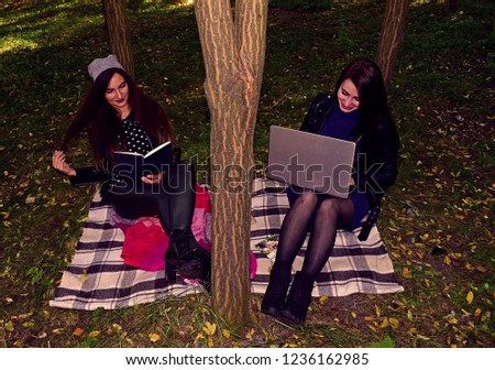 Two girls with a laptop and a book sitting in the city Park near the trees
