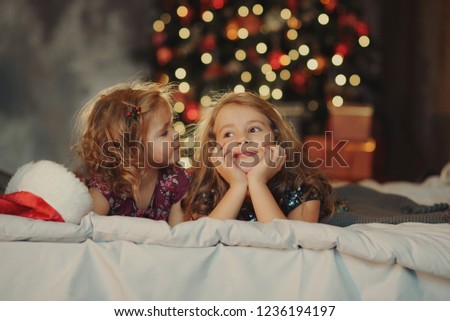 Two  cute sisters in Santa Claus hats are sitting on a soft home bad against the background of a festive Christmas tree  hugging and smiling. Home Family New Year's holiday. Children's relationships