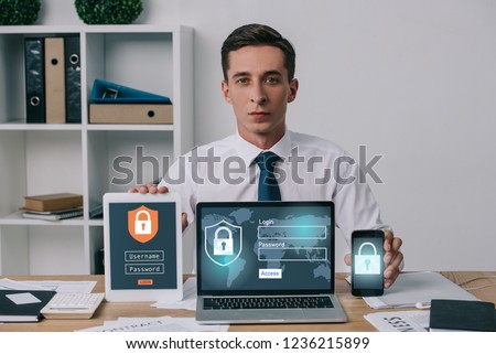 portrait of businessman showing laptop, tablet and smartphone with cyber security signs on screens at workplace in office