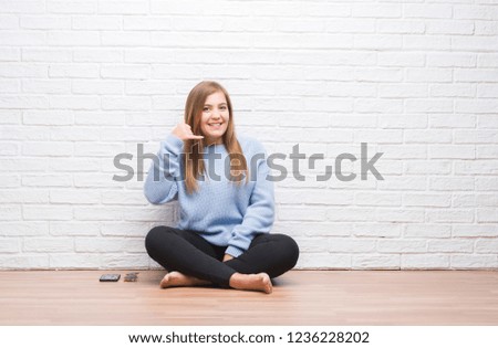 Young adult woman sitting on the floor in autumn over white brick wall smiling doing phone gesture with hand and fingers like talking on the telephone. Communicating concepts.