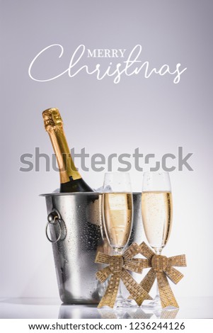 bottle of champagne in bucket and glasses of champagne on grey backdrop with "merry christmas" lettering