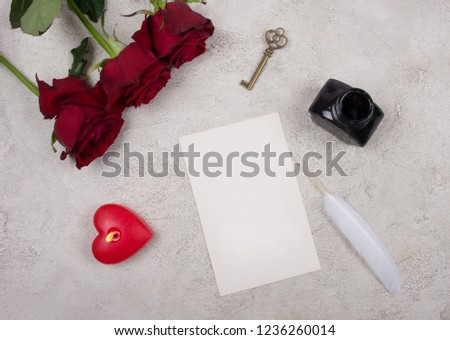 Red roses, a heart-shaped candle, a key, a blank card, a white feather and an inkpot on a gray marble or concrete background (top view) as the Valentine day or love concept