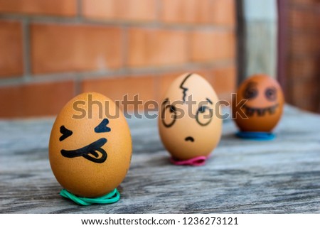 Three eggs with funny faces drawn on them are standing on the woodern chair in front of a brick wall. It's summer