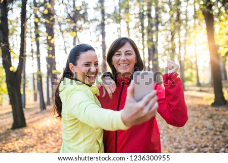 Two female runners with smartphone taking selfie outdoors in forest in autumn nature.