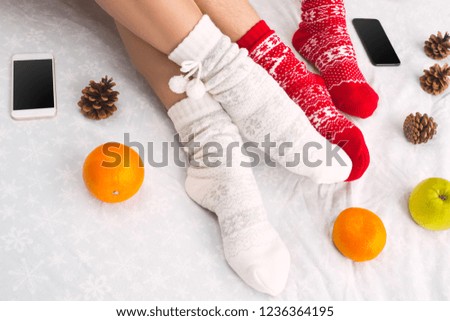 Soft photo of woman and man on the bed with phone and fruits, top view point. Female and male legs of couple in warm woolen socks. Christmas, love, lifestyle concept