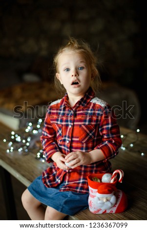 A beautiful little blonde in a traditional plaid shirt opens a chocolate bar from her sweet Christmas gift, sitting on a wooden table with a garland in the background. Christmas morning concept.