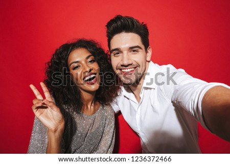 Portrait of a cheerful young smartly dressed couple standing isolated over red background, taking a selfie with outstretched hand