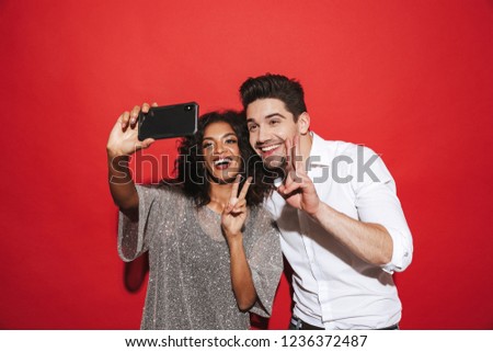 Portrait of a cheerful young smartly dressed couple standing isolated over red background, taking a selfie