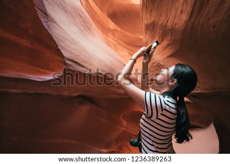 young asian lady tourist standing in the lower antelope canyon. beautiful traveler holding cellphone using photo app to take picture of the amazing desert view from down place. pretty girl join tour