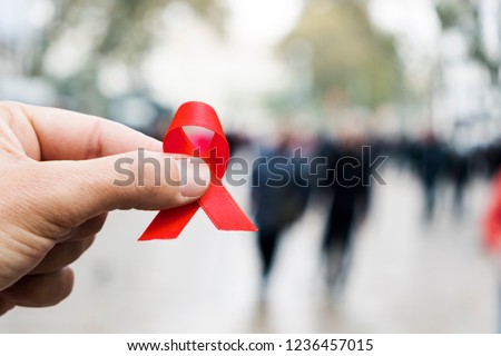 closeup of a red awareness ribbon for the fight against AIDS in the hand of a young caucasian man in a busy pedestrian street of a city Royalty-Free Stock Photo #1236457015