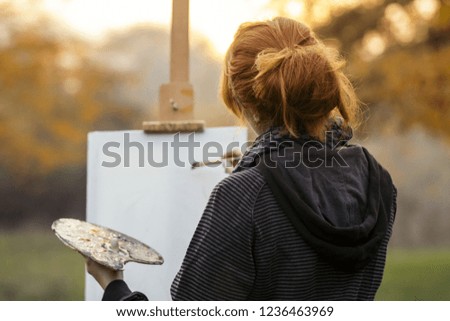 red-haired girl with a bun painting a picture on an easel in nature, a young woman involved in creativity and enjoying beautiful landscape at sunset , concept of seasons mood and hobby