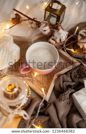 Mug of hot cappuccino on a wooden tray is on the bed. Cozy decor. Breakfast. Mug, plaid, cotton, candle. Gift box and knitted mittens. Christmas lights. Holidays.