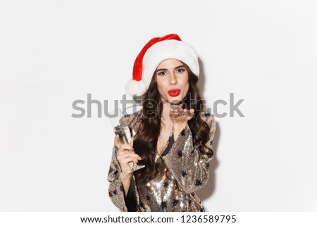 Happy young woman wearing shiny dress and santa hat celebrating New Year isolated over white background, holding glass of champagne, send kiss