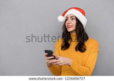 Portrait of a smiling young woman wearing red santa claus hat standing isolated over gray background, using mobile phone