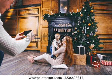 Theme mobile photography, amateur photo video phone. Hands Caucasian man holds uses smartphone makes photo mother and son home near fireplace and Christmas tree winter Christmas time New Year decor.