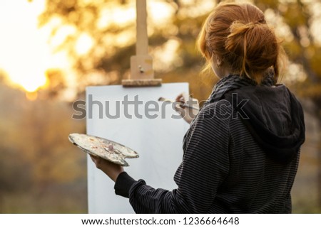 red-haired girl with a bun painting a picture on an easel in nature, a young woman involved in creativity and enjoying beautiful landscape at sunset , concept of seasons mood and hobby