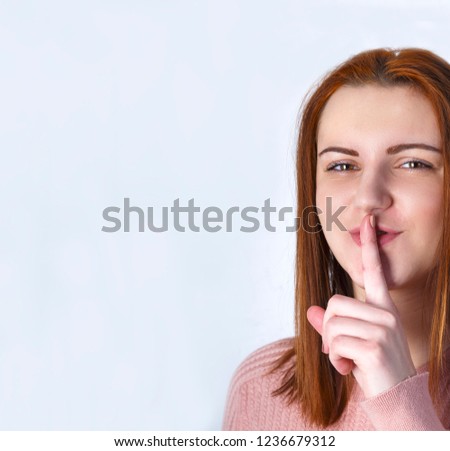 Young sly smiling redhead girl with pink sweatshirt showing sign of closing mouth and silence gesture putting finger in mouth