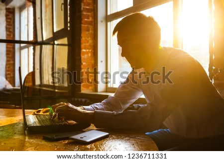 Young pensive coworker working at sunny work place loft while sitting at wooden table.Man analyze document on laptop display.Blurred background