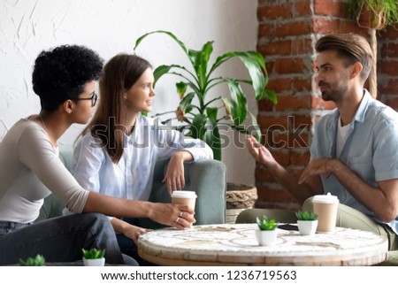 Diverse friends meeting in coffee house talking at table, friendly man telling story to women, gesticulate, workmates or colleagues spending time together in cafeteria during break, enjoying coffee