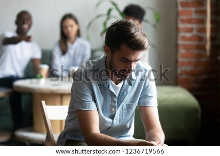Upset frustrated young man suffering from gossiping, bullying, discrimination, avoiding, ignoring, having problem with bad friends, feeling offended and hurt, sitting alone in cafe Royalty-Free Stock Photo #1236719566