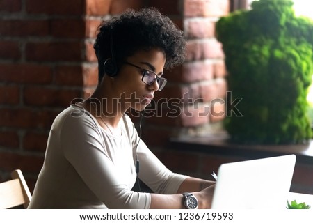 Focused African American woman wearing headphones using laptop, making writing notes, serious female student learning language, watching online webinar, listening audio course, e-learning, education