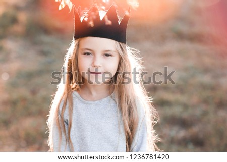 Smiling blonde baby girl 4-5 year old wearing handmade crown outdoors. Looking at camera. Childhood. 
