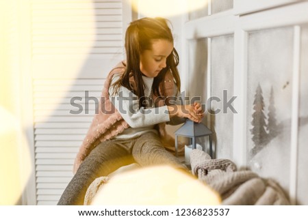 Little girl sitting by the window with a cup of hot drink and looking at the winter forest