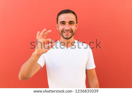 Picture of happy young handsome man standing isolated over red wall background. Looking camera showing okay gesture.