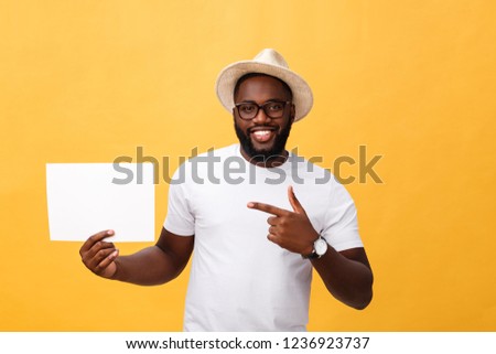 Picture of young smiling african-american man holding white blank board and pointing on it, on yellow background, copy space
