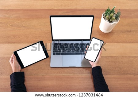 Top view mockup image of a hands holding mobile phone , black tablet and laptop with blank white screen on wooden table in office