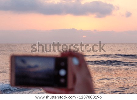 Girl's hand holding smart phone taking sunset photo on the beach. Mobile phone with sunset view