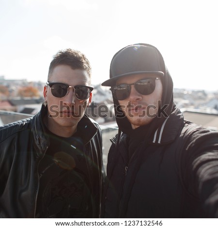 Two cool fashionable young man friends with fashion sunglasses in black fashionable clothes with a cap and a hood make selfie against the background of the city
