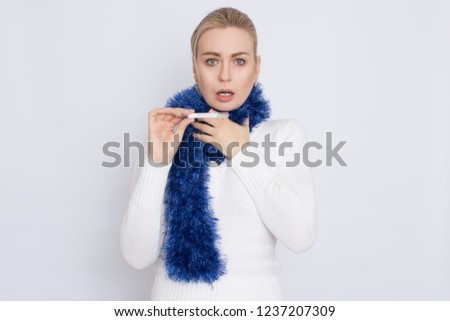 Portrait of sad sick blonde woman dressed in sweater and scarf over white background, showing thermometer