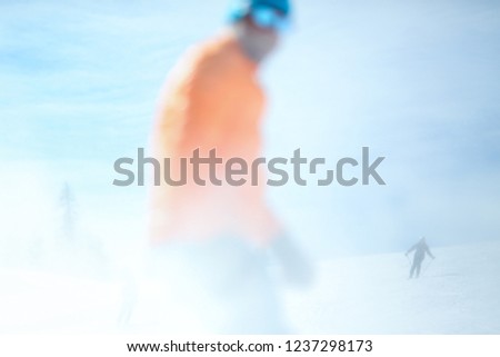 Conceptual photography. A male skier in an orange jacket is out of focus in the foreground. In the background rides a skier