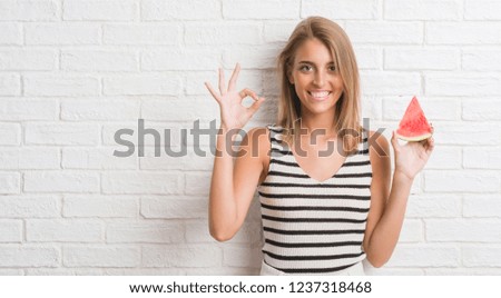 Beautiful young woman over white brick wall eating water melon slice doing ok sign with fingers, excellent symbol