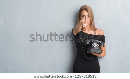 Beautiful young woman over grunge grey wall holding vintage telephone pointing with finger to the camera and to you, hand sign, positive and confident gesture from the front