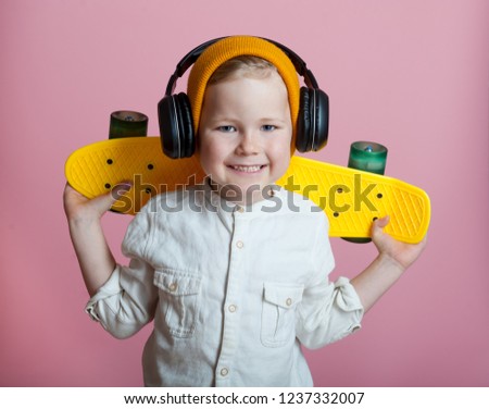 Stylish little school boy with skateboard and earphones and wearing fashion yellow hat and glasses on a colorful pink background. Happy smiling child with longboard.