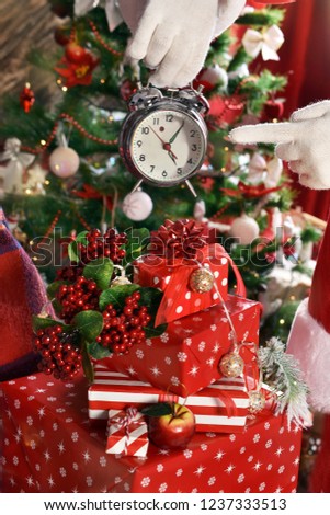 santa claus  points his finger at alarm clock above the stack of gifts in front of christmas tree 