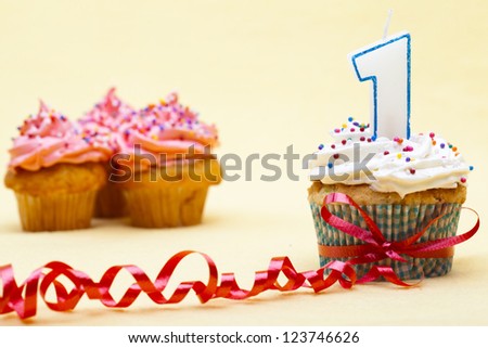 Close-up image of a cupcake with number 1 candle and red streamer while strawberry cupcake in background.