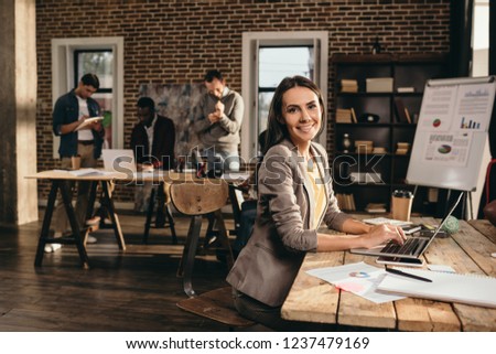 smiling business woman sitting at desk with laptop and working on project at loft office with colleagues on background