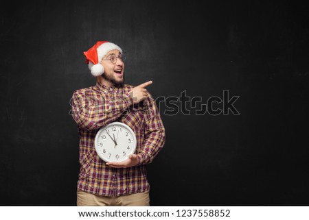 Christmas men with clock. Portrait of surprised young man wearing red santa claus hat, holding clock in hands, on black background. Fun emotion facial expression. Last minute christmas shopping