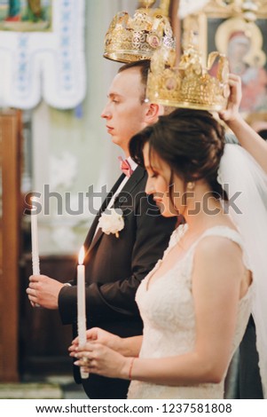 Happy stylish wedding couple holding candles with light under golden crowns during holy matrimony in church. Bride and groom standing at wedding ceremony