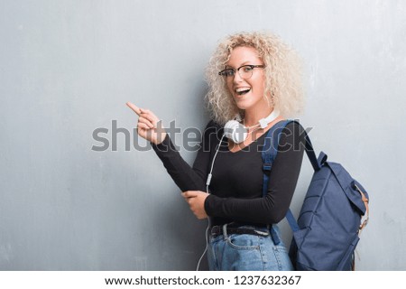 Young blonde woman over grunge grey wall wearing backpack and headphones with a big smile on face, pointing with hand and finger to the side looking at the camera.