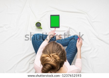 Woman uses a tablet, sitting on a white crumpled blanket, green Cup of coffee or tea, women's feet. Women's hands. Background with copy space, for advertisement. Top view